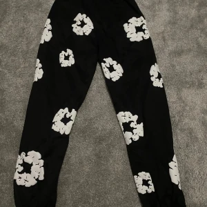 Svarta Denim Tears mjukisbyxor med tryck - Svarta mjukisbyxor från Denim Tears med vita blommiga tryck över hela benen. Byxorna har resår vid bensluten och midjan för skön passform. Perfekt för chill dagar eller streetwear-stil. Materialet är mjukt och bekvämt, och designen sticker ut med sitt unika mönster.