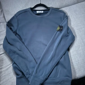 Mörkblå sweatshirt från Stone Island - Säljer en stilren mörkblå sweatshirt från Stone Island storlek L passar M med klassisk rund hals och ikonisk logga på ärmen. Tröjan har långa ärmar och är tillverkad i mjukt material som känns skönt mot huden. Perfekt för dig som gillar streetwear och enkel design.
