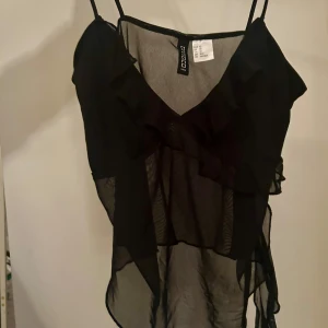 Svart volangtop från H&M - Säljer en svart, transparent linnetopp från Divided med tunna axelband och snygga volangdetaljer framtill. Toppen har v-ringning och är tillverkad i ett lätt, luftig material som ger en cool och trendig look.