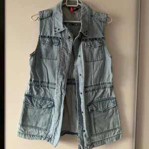 Ljusblå jeansväst med fickor från H&M - Snygg ljusblå jeansväst från H&M med fyra stora fickor framtill och tryckknappar. Västen har klassisk krage och markerad midja med resårdetalj. Perfekt att slänga över en hoodie eller t-shirt för en chill look.