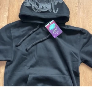 Svart hoodie från SAIK med tryck - Svart hoodie från SAIK med stort tryck på huvan och grova snören. Klassisk känguruficka framtill och mjukt material som känns skönt mot huden. Perfekt för dig som gillar streetwear och vill ha en stilren look.