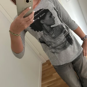 Grå långärmad topp med tryck  - Grå topp från Reserved i storlek XS. Snyggt svart tryck framtill med text och porträtt, rund halsringning och mjukt material. Perfekt till jeans eller vida byxor för en avslappnad look.
