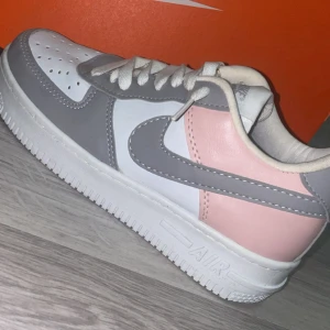 Nike Air Force 1 i grått, vitt och rosa - Snygga Nike Air Force 1 sneakers i en fräsch färgkombo av grått, vitt och ljusrosa. Klassisk låg modell med vit sula, grå swoosh och detaljer, samt rosa panel på hälen. Perfekta för dig som vill sticka ut med stilrena sneakers.