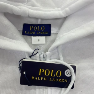 Vit hoodie från Polo Ralph Lauren S - Vit hoodie från Polo Ralph Lauren med dragkedja framtill och klassisk röd broderad logga på bröstet. Tröjan har huva med snörning, kängurufickor och långa ärmar. Perfekt för en clean och stilren look.