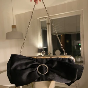 Svart kuvertväska med strassdetalj - Stilren svart kuvertväska i satin med silverfärgad kedja och ram och en stor rosett med en cirkel av glittrande strass på framsidan🤍 perfekt för en utekväll med det allra viktigaste!! I nytt skick!