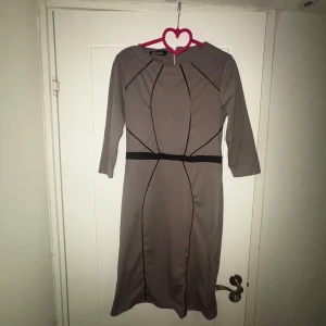 Beige klänning med svarta detaljer - Snygg beige långklänning från Exclusiv med svarta linjer som ger en cool grafisk look. Klänningen har trekvartsärm och markerad midja med svart band. Perfekt för dig som gillar stilrena och moderna plagg.