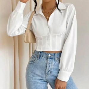 Vit croppad skjorta med snörning - Snygg vit croppad skjorta med långa ärmar och markerad midja. Skjortan har klassisk krage, knappar framtill och cool snörning i ryggen som ger en unik look. Perfekt att styla med jeans för en trendig outfit.