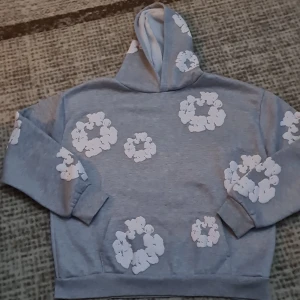 Grå hoodie från Denim Tears med blommor - Säljer en grå hoodie från Denim Tears med vita blommiga tryck över hela tröjan. Hoodien har huva, långa ärmar och en relaxed passform. Perfekt för dig som gillar streetwear och vill sticka ut med en unik design.