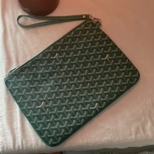 Grön Goy4rd kuvertväska - Säljer en grön kuvertväska från goy4rd med ett ikoniskt grafiskt mönster och silverfärgad dragkedja. Väskan har avtagbar handledsrem och insida i orange textil. Perfekt för att bära det viktigaste och ge din outfit en lyxig touch.