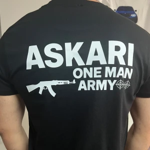Svart ASKARI t-shirt med tryck - Svart t-shirt från ASKARI med vit text och grafik på både bröstet och ryggen. Trycket har texten 'ASKARI ONE MAN ARMY' och en AK-47 illustration samt måltavlor. T-shirten har rund hals och korta ärmar, perfekt för dig som gillar streetwear med attityd.