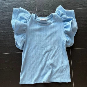 Blå ribbad topp med volangärmar Zara - Supersöt ljusblå topp från Zara med ribbad struktur och stora volangärmar som verkligen sticker ut. Rund halsringning och figurnära passform. Perfekt för dig som vill ha något extra till din outfit!
