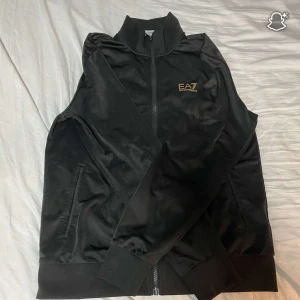 Svart track suit från EA7 Armani - Svart track suit från EA7 Emporio Armani med guldfärgad logga på bröstet. Tröjan har hög krage, dragkedja framtill och ribbade muddar vid ärmslut och nederkant. Tillverkad i glansig polyester för en sportig vibe.