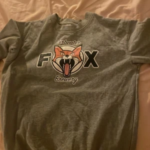 Grå sweatshirt FOX Dirtwater Brewery - Grå sweatshirt med tryck av en räv och texten 'FOX Dirtwater Brewery' på bröstet. Tröjan har ribbade muddar vid ärmslut och nederkant samt rund halsringning.