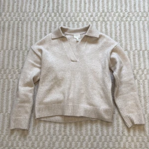 Beige stickad tröja med krage - Beige stickad tröja från H&M i storlek XS. Tröjan har en snygg krage och v-ringning, samt ribbade muddar vid ärmslut och nederkant. Perfekt för lager-på-lager och chill höst- eller vinterstil.