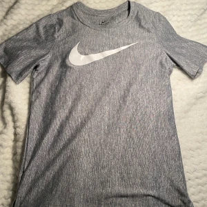 Grå nike tränings t-shirt - Snygg gråmelerad t-shirt från Nike med stor vit logga på bröstet. Tillverkad i Dri-Fit polyester som andas och håller dig torr. T-shirten har rund hals, korta ärmar och en normal passform. Perfekt för träning eller chill. Oanvänd men ingen prislapp kvar 