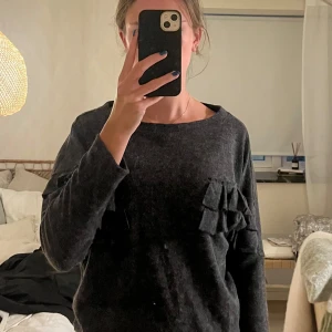 Mörkgrå volangtröja Holly Bracken - Mysig mörkgrå tröja från Holly Bracken med volangdetaljer på axlarna. Tröjan har rund halsringning och långa ärmar. Tillverkad i en mjuk blandning av polyester, viskos och akryl. Perfekt för dig som gillar en chill men ändå trendig look.