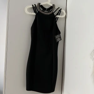 Svart strappy klänning från NLY ONE - Supersnygg svart kortklänning från NLY ONE med flera smala axelband och dekorerad halsringning med pärlor och glittriga detaljer. Klänningen har en tight passform och hög hals, perfekt för dig som vill sticka ut. Materialet är stretchigt och känns lyxigt. På grund av strul med bankkonto så uppskattas kontanter. Alltså kommer det behövas att hämta paketet.