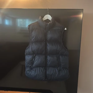 Svart dunväst med dragkedja - Snygg svart dunväst med hög krage och två praktiska fickor med dragkedja på framsidan. Västen har en quiltad design och stängs med en hel dragkedja. Perfekt att slänga över en hoodie för extra värme när det är kyligt ute.