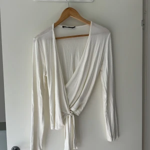 Vit omlottopp från Lager 157, stl M - Snygg vit omlottopp från Lager 157 i mjukt material. Toppen har långa ärmar, djup v-ringning och knyt i midjan för en trendig look. Perfekt att styla med jeans eller kjol för en chill vibe.