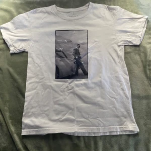 Vit t-shirt med svartvit print - Säljer en vit t-shirt i storlek S med en stor svartvit print på framsidan som visar en person lutad mot en bil med berg i bakgrunden. T-shirten har rund hals och korta ärmar, tillverkad i mjuk bomull. Perfekt för dig som gillar coola motiv.