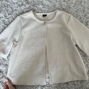 Beige cardigan bolero kofta från Gina Tricot - Stilren beige cardigan bolero kofta från Gina Tricot med rund halsringning och en snygg guldig knapp framtill. Jackan har trekvartsärmar och är tillverkad i ett mjukt material som känns skönt mot huden. Perfekt för dig som gillar minimalistisk och clean stil. Passar xs, s o m