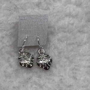 Söta silverörhängen med jordgubbar - Charmiga örhängen i silverfärgad metall med små hängande jordgubbar som motiv. Perfekta för dig som vill ha en lekfull och unik detalj till din stil. Lätta att bära och passar till alla tillfällen. Krokarna är i sterling silver 