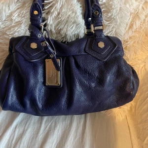 Mörkblå skinnväska Marc by Marc Jacobs - Säljer en snygg mörkblå handväska från Marc by Marc Jacobs i äkta skinn. Väskan har silverfärgade metalldetaljer, dubbla handtag, avtagbar axelrem och dragkedja upptill. Insidan har coolt tryckt foder med logga. Perfekt för dig som vill ha en stilren och rymlig väska.