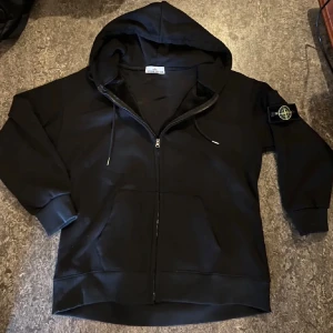 Svart hoodie från Stone Island - Svart hoodie från Stone Island med dragkedja framtill, huva med snörning och klassisk logotyp-patch på vänster ärm. Tröjan har känguruficka och är tillverkad i mjuk bomull. Perfekt för en chill och stilren look.