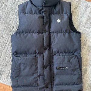 Svart dunväst från Canada Goose XS - Säljer en svart dunväst från Canada Goose i storlek XS. Västen har hög krage, stora framfickor och knäppning med svarta knappar. Klassisk Canada Goose-logga på bröstet och etikett på fickan. Perfekt för kalla dagar när du vill hålla stilen.