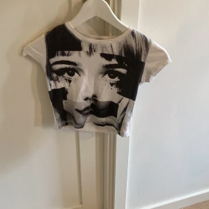 Vit croppad topp med ansikte Lindex - Cool croppad topp från Lindex med stort svartvitt ansikte tryckt på framsidan. T-shirten är vit med korta ärmar och har en riktigt edgy vibe. Perfekt för dig som vill sticka ut med ett unikt print. Gjord i mjuk bomull.