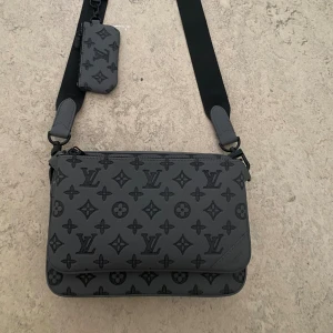 Grå Louis Vuitton axelväska med monogram - Säljer en grå axelväska från Louis Vuitton med klassiskt monogrammönster i svart. Väskan har justerbar axelrem, två fack med dragkedja och en liten matchande plånbok. Tillverkad i skinn med snygga detaljer och stilren design. Säljer väskan på grund av jag inte hinner använda den jobbar 6 dagar suckas även med kvitto 