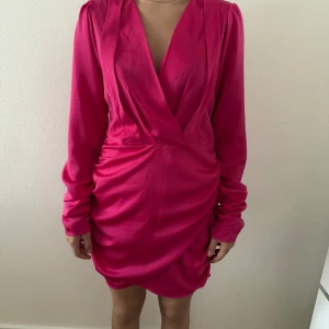 Rosa omlottklänning med v-ringning - Snygg kort rosa klänning med omlott och djup v-ringning. Klänningen har långa ärmar och draperad effekt både fram och bak, vilket ger en cool och trendig look. Perfekt för dig som vill sticka ut med en färgstark outfit.