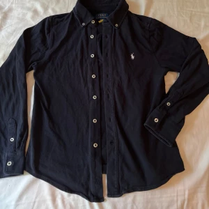 Mörkblå skjorta från Polo Ralph Lauren - Klassisk mörk blå skjorta från Polo Ralph Lauren med vit broderad logga på bröstet. Skjortan har lång ärm, knappar hela vägen och en snygg krage. Tillverkad i mjuk bomull som känns skön mot huden. Perfekt för dig som gillar stilrena och tidlösa plagg. Kontakta om ni har frågor och priset är inte ristat i sten. Org pris 1000kr