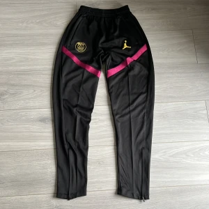 PSG x Jordan svarta träningsbyxor - Svarta träningsbyxor från PSG x Jordan med gult broderat PSG-märke och Jumpman-logga. Snygga lila detaljer över låren, smal passform och dragkedja vid benslut. Perfekta för fotboll eller chill. Tillverkade i polyester för skön känsla.