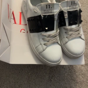 Valentino Rockstud Untitled sneakers - Säljer ett par vita Valentino Rockstud Untitled sneakers med coola svarta detaljer och nitar över snörningen. Skorna har rund tå, platt sula och är tillverkade i skinn med vita snören. Perfekt för dig som vill sticka ut med en lyxig och edgy look. ‼️TRE SPIKAR HAR GÅTT AV DÄRAV PRISET‼️