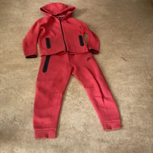 Röd Nike träningsset med huva - Nike träningsset i rött med svarta detaljer. Setet består av en hoodie med dragkedja och två svarta fickor samt matchande byxor med svarta fickor och Nike-logga. Mjukt material, perfekt för sport eller chill. Snygg sportig look med ribbade muddar. Är bara använd ett fåtal gånger