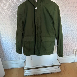 Grön jacka från Superdry  st S - Snygg grön jacka  från Superdry International Atlantic Race med huva och stora fickor framtill. Jackan har coola logodetaljer på ärmen och insidan, samt vita knappar. Perfekt för dig som gillar sportig och clean stil.  Träffa i Gbg 