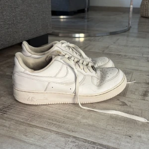 Nike Air Force 1 vita sneakers - Klassiska Nike Air Force 1 sneakers i helvitt skinn med lågt skaft. Skorna har rund tå, perforerad tåbox och vit sula med tydlig Air-dämpning. Perfekta för en clean och tidlös streetstil. Ikonisk Swoosh-logga på sidorna och snörning framtill.