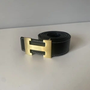 Svart skärp med H-spänne - Snyggt svart skärp från H-ermes i äkta skinn med ikoniskt H-format spänne i guldmetall. Bältet har vita kontrastsömmar längs kanterna och en stilren, klassisk design som lyfter vilken outfit som helst.