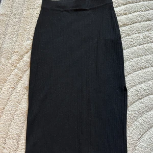 Svart ribbad långkjol från ONLY - Svart ribbad långkjol från ONLY i figurnära modell med snygg slits på sidan. Kjolen är stretchig och har en enkel, stilren look som passar perfekt till crop tops eller oversized tröjor. Materialet är mjukt och bekvämt.