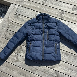 Mörkblå pufferjacka från JH Collection - Vinterjacka till stallet. Säljer en mörkblå pufferjacka från JH Collection med hög krage och tre dragkedjeförsedda fickor. Jackan har quiltad design och är perfekt för kyligare dagar. Materialet är syntet och jackan är långärmad med en normal passform.
