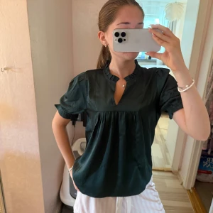 Mörkgrön blus med kort ärm - Säljer en mörkgrön blus med korta puffärmar och en liten ståkrage med v-ringad öppning. Blusen har en luftig passform och fina rynkdetaljer under bysten. Perfekt för dig som gillar stilrena och bekväma toppar.