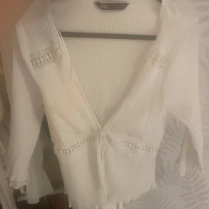 Vit omlottblus från Zara - Superfin vit omlottblus från Zara med v-ringning, knyt i midjan och dekorativa spetsdetaljer på ärmarna. Blusen har en lätt skrynklig struktur och trekvartsärmar, perfekt för en fräsch och bohemisk look.