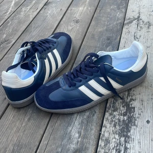Adidas Samba sneakers marinblå pris kan sänkas - Säljer ett par Adidas Samba sneakers i marinblått och vitt med klassiska tre ränder på sidan. Skorna har snörning, platt sula och rund tå. Ovandelen är i skinn och mocka, med vit häl och grå yttersula. Perfekt för dig som gillar retrostil och sportig vibe.