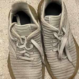 Beige Adidas sneakers strl 38 2/3 - Säljer ett par beige Adidas sneakers med stickad ovandel och detaljer i skinn. Skorna har snörning och en chunky, mönstrad gummisula i gulbrun ton. Perfekta för dig som gillar en sportig och modern look.