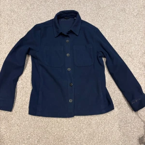 Mörkblå overshirt från d’once de la sense - Snygg mörkblå overshirt. Med två stora bröstfickor och klassisk krage. Jackan har knappar hela vägen och broderad logga på ärmen. Materialet är mjukt och känns som ull, perfekt för lager-på-lager. Passformen är normal och ärmarna är långa.