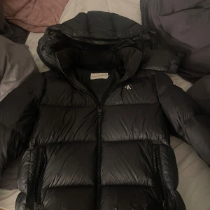 Svart pufferjacka Calvin Klein Jeans -  Säljer min calvin klein jacka då den inte längre använd, nyrpis 1600kr. Den är i bra skick förutom att den fick ett hål (bild 4) som jag tejpat med special tejp över.