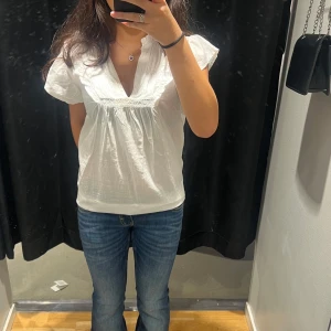 Vit blus med volangkrage - Söt vit blus med korta ärmar och volangkrage. Blusen har v-ringning och fina rynkade detaljer framtill. Tillverkad i ett lätt och luftigt bomullstyg, perfekt för varmare dagar.