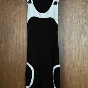 Svart och vit retro kortklänning - Cool svart och vit kortklänning med breda axelband och stora knappar framtill. Klänningen har kontrasterande vita detaljer vid axelpartiet och fickorna, vilket ger en retrovibe. Materialet känns mjukt och stretchigt, perfekt för dig som gillar en unik look.