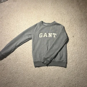 Grå sweatshirt från GANT - Klassisk grå sweatshirt från GANT med broderad logga framtill i vitt. Tröjan har rund hals, ribbade muddar och är tillverkad i mjuk bomullsblandning. Perfekt för en chill och stilren look. Använd 1 gång💕 